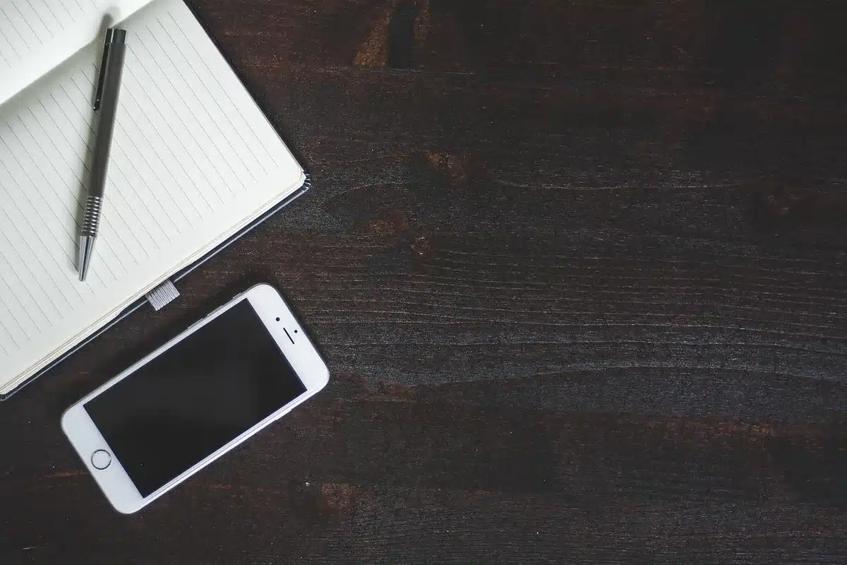 A small, unbranded compact smartphone resting on a wooden table beside a closed notebook and a coffee cup, screen off, suggesting everyday use and longevity