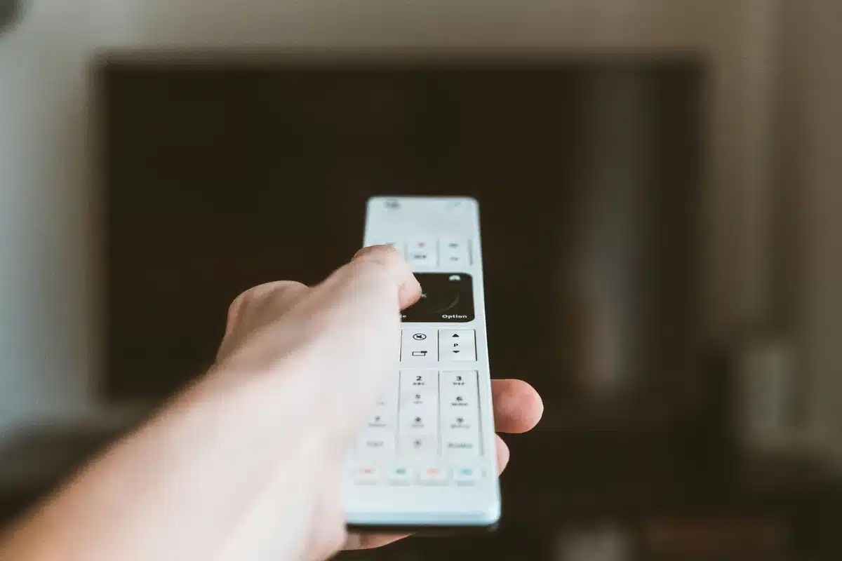 Close view of an unbranded remote control lying on a table with a blurred large flat-screen TV in the background, anonymous hands unreachable; clean modern interior, no logos